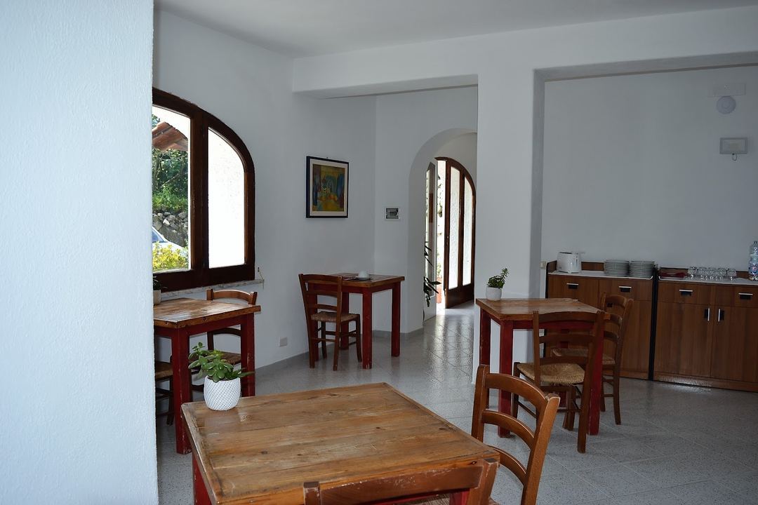 A bright, open-plan dining and kitchen area with wooden tables, chairs, and rustic furnishings, featuring arched doorways and white walls.