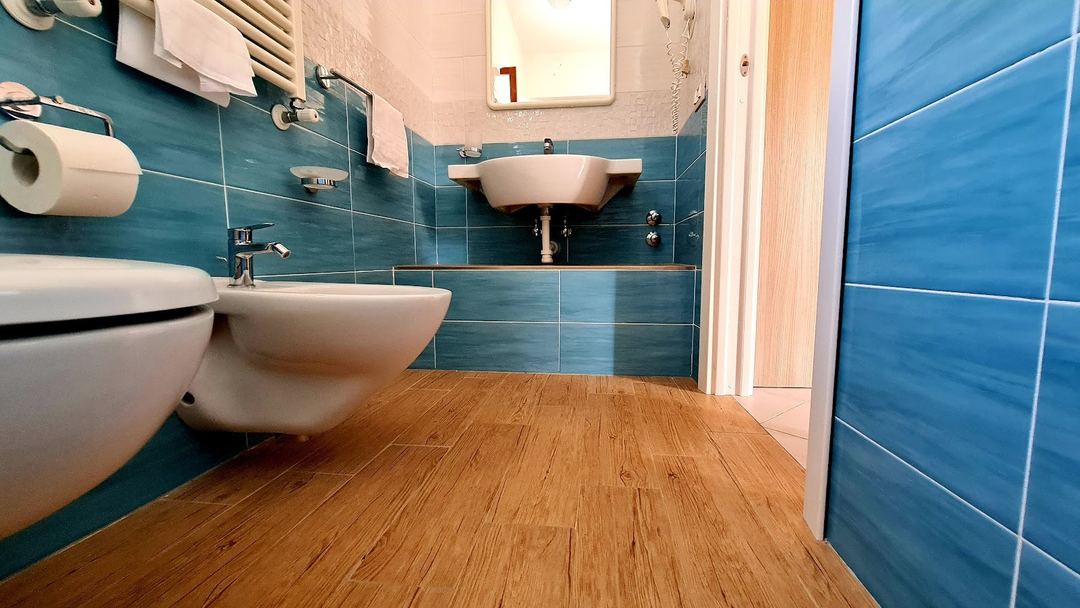 A modern bathroom features teal blue tile walls, wooden flooring, and white ceramic fixtures including a toilet and two wall-mounted sinks.