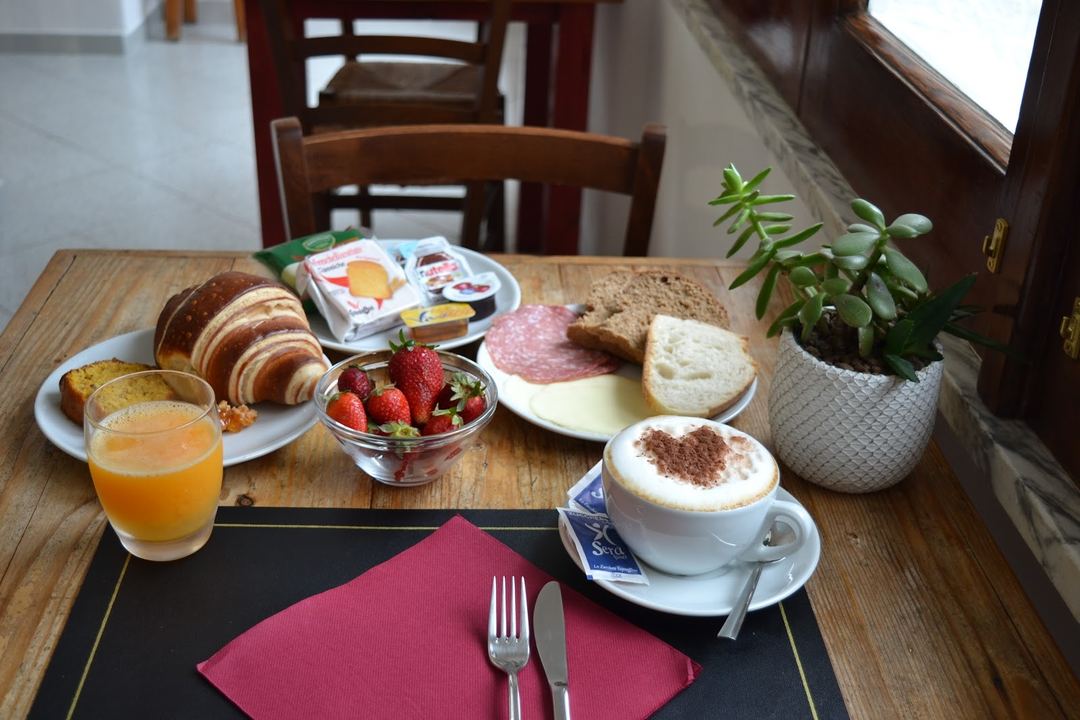 A beautifully arranged breakfast spread featuring croissants, fresh strawberries, cured meats, cheese, bread, orange juice, and cappuccino on a wooden table with place settings.